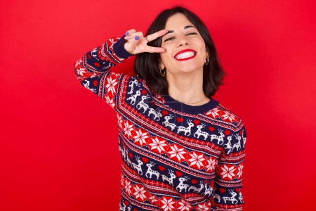 Téléchargez les photos : Brune caucasienne portant chandail de Noël sur fond rouge Faire symbole de paix avec les doigts sur le visage, souriant gai montrant la victoire - en image libre de droit