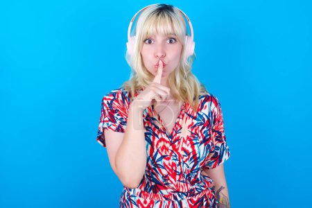 Photo for Caucasian girl wearing floral dress isolated over blue background making hush gesture with finger on her lips wearing  wireless headphones. Be quiet. - Royalty Free Image