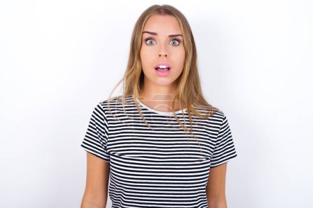 Photo for Shocked beautiful blonde girl wearing striped t-shirt on white background stares bugged eyes keeps mouth opened has surprised expression. Omg concept - Royalty Free Image