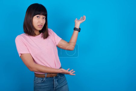 Téléchargez les photos : Jeune femme asiatique portant t-shirt sur fond bleu pointant de côté avec les deux mains montrant quelque chose d'étrange et disant : Je ne sais pas ce que c'est. Concept de publicité. - en image libre de droit