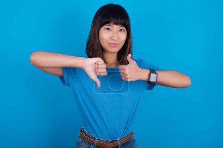 Foto de Joven asiático mujer usando t-shirt contra azul fondo mostrando pulgar arriba abajo signo - Imagen libre de derechos