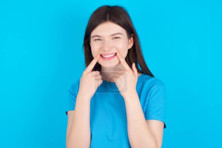 Téléchargez les photos : Heureuse jeune fille caucasienne portant T-shirt bleu isolé sur fond bleu avec sourire aux dents, garde les index près de la bouche, les doigts pointant et forçant sourire joyeux - en image libre de droit