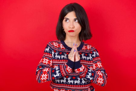Photo for Charming cheerful brunette caucasian woman wearing christmas sweater over red background making up plan in mind holding hands together, setting up an idea. Look askance - Royalty Free Image