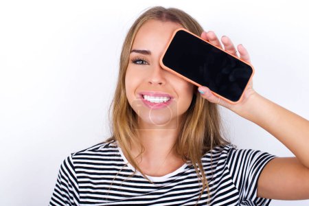 Foto de Hermosa chica rubia con camiseta a rayas sobre fondo blanco sosteniendo teléfono inteligente moderno que cubre un ojo mientras sonríe - Imagen libre de derechos