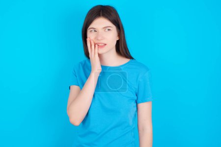 Téléchargez les photos : Concept de mal de dents. Plan intérieur d'une jeune fille caucasienne portant un T-shirt bleu isolé sur fond bleu ressentant de la douleur, tenant sa joue avec la main, souffrant de mal de dents, regardant la caméra avec une expression douloureuse - en image libre de droit