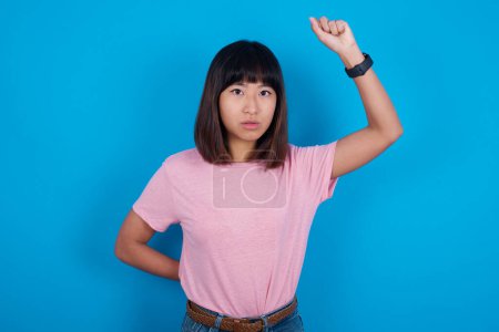 Photo for Young asian woman wearing t-shirt against blue background feeling serious, strong and rebellious, raising fist up, protesting or fighting for revolution. - Royalty Free Image