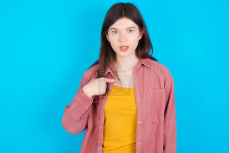 Photo for Young caucasian girl wearing pink shirt isolated over blue background being in stupor shocked, has astonished expression pointing at oneself with finger saying: Who me? - Royalty Free Image