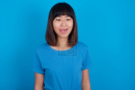 Téléchargez les photos : Drôle jeune asiatique femme porter t-shirt sur bleu fond fait grimace et croix yeux joue fou a amusant seul bâtons sur la langue. - en image libre de droit