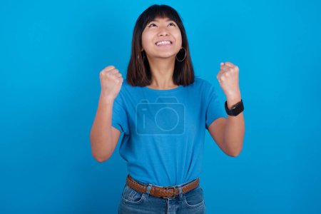 Téléchargez les photos : Jeune femme asiatique portant t-shirt sur fond bleu. Jeune champion ayant du succès être heureux d'atteindre ses objectifs. Concept de victoire et de triomphe. - en image libre de droit