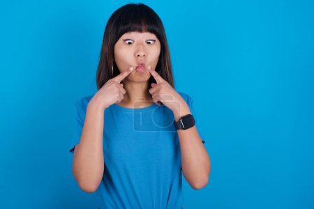 Téléchargez les photos : Jeune femme asiatique portant t-shirt sur fond bleu croise les yeux et rend les lèvres de poisson grimace drôle - en image libre de droit