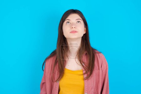 Téléchargez les photos : Jeune fille caucasienne portant chemise rose isolé sur fond bleu levant les yeux comme il voit quelque chose d'étrange. - en image libre de droit