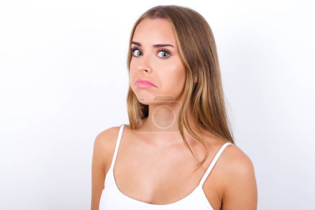 Young Caucasian girl wearing white tank top on white background with snobbish expression curving lips and raising eyebrows, looking with doubtful and skeptical expression, suspect and doubt.