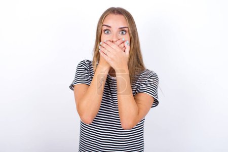Stunned beautiful blonde girl wearing striped t-shirt on white background covers mouth with both hands being afraid from something or after hearing stunning gossips.