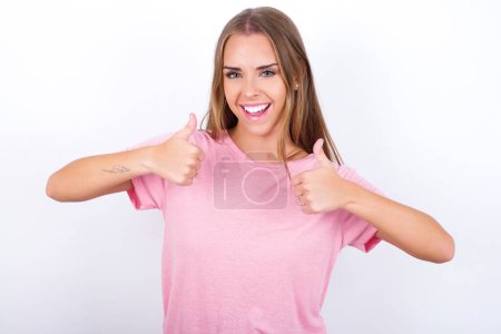 Foto de Optimistic Joven Caucásico niña con una camiseta rosa sobre fondo blanco que muestra los pulgares hacia arriba con emociones positivas. Concepto de calidad y recomendación. - Imagen libre de derechos