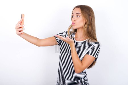 Photo for Beautiful blonde girl wearing striped t-shirt on white background blows air kiss at camera of smartphone and takes selfie, sends mwah via online call. - Royalty Free Image