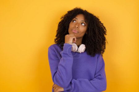 Téléchargez les photos : Portrait de jeune femme afro-américaine réfléchie avec écouteurs sur fond jaune - en image libre de droit