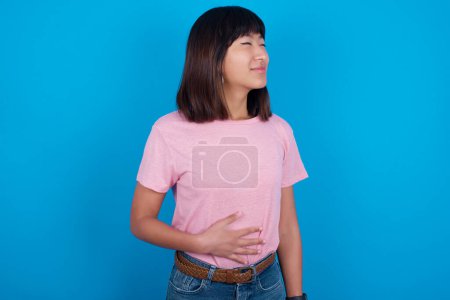 Téléchargez les photos : Satisfait souriant jeune femme asiatique portant t-shirt sur fond bleu garde les mains sur le ventre, être de bonne humeur après avoir mangé un délicieux souper, démontre qu'elle est pleine. Sensation agréable dans l'estomac. - en image libre de droit