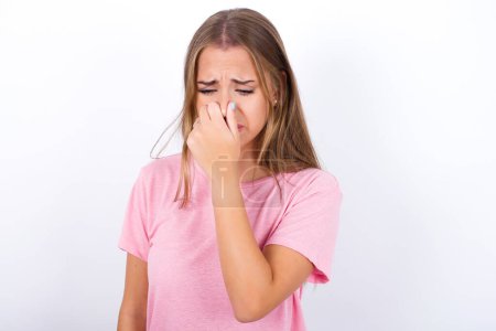 Foto de Joven chica caucásica con camiseta rosa sobre fondo blanco sosteniendo su nariz debido a un mal olor. - Imagen libre de derechos