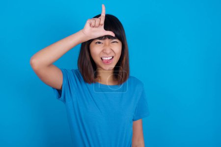 Téléchargez les photos : Jeune femme asiatique portant t-shirt sur fond bleu gestes avec le doigt sur le front fait geste perdant se moque des gens montre la langue - en image libre de droit