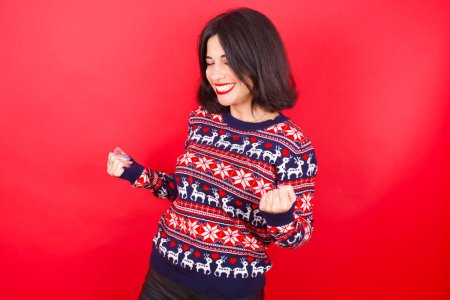 Téléchargez les photos : Brune caucasienne portant chandail de Noël sur fond rouge très heureux et excité faire geste gagnant avec les bras levés, souriant et criant pour le succès. Concept de célébration. - en image libre de droit