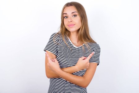Photo for Beautiful blonde girl wearing striped t-shirt on white background crosses arms and points at different sides hesitates between two items or variants. Needs help with decision - Royalty Free Image