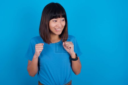 Foto de Joven asiático mujer usando t-shirt contra azul fondo aprieta puños y espera para algo agradable sucedió mira lejos muerde labios y espera anuncio de resultados - Imagen libre de derechos