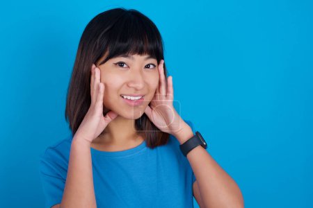 Foto de Feliz joven asiática mujer usando camiseta contra fondo azul toca ambas mejillas suavemente, tiene sonrisa tierna, muestra dientes blancos, mira positivamente directamente a la cámara, - Imagen libre de derechos