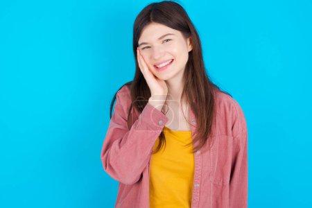 Photo for Shocked, astonished young caucasian girl wearing pink shirt isolated over blue background looking surprised in full disbelief wide open mouth with hand near face. Positive emotion facial expression body language. - Royalty Free Image