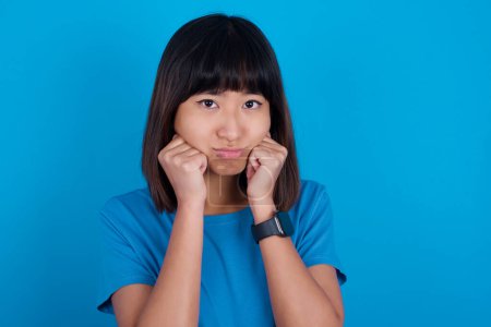 Photo for Portrait of sad young asian woman wearing t-shirt against blue background hands face - Royalty Free Image