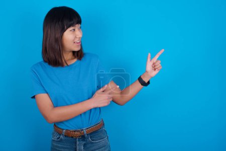 Foto de Joven asiático mujer usando t-shirt contra azul fondo sonrisa excitado dirigiendo dedos mirar vacío espacio - Imagen libre de derechos