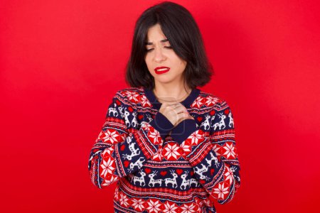 Téléchargez les photos : Triste femme caucasienne brune portant un pull de Noël sur fond rouge désespérée et déprimée avec des larmes sur les yeux souffrant de douleur et de dépression dans la tristesse expression faciale et concept d'émotion - en image libre de droit