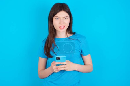 Foto de Foto de chica caucásica loca asombrada usando camiseta azul aislada sobre fondo azul mantenga el concepto de retroalimentación de disgusto del teléfono inteligente - Imagen libre de derechos
