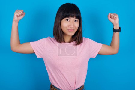 Foto de Cintura arriba tiro de joven mujer asiática usando camiseta contra fondo azul levanta los brazos para mostrar los músculos se siente seguro de la victoria, se ve fuerte e independiente, sonríe positivamente a la cámara. Concepto deportivo. - Imagen libre de derechos