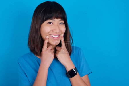Foto de Feliz joven asiática mujer usando camiseta sobre fondo azul con sonrisa dentada, mantiene los dedos índice cerca de la boca, dedos señalando y forzando sonrisa alegre - Imagen libre de derechos
