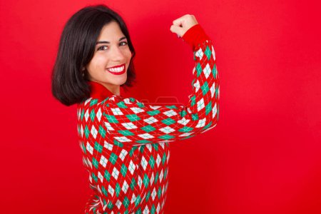 Téléchargez les photos : Portrait de puissante femme caucasienne brune joyeuse portant un pull de Noël sur fond rouge montrant les muscles. - en image libre de droit