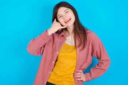 Photo for Young caucasian girl wearing pink shirt isolated over blue background imitates telephone conversation, makes phone call gesture with hands, has confident expression. Call me! - Royalty Free Image