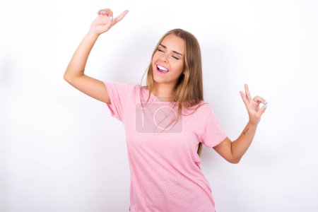 Photo for Photo of upbeat Young Caucasian girl wearing pink T-shirt on white background has fun and dances carefree wear being in perfect mood makes movements. Spends free time on disco party - Royalty Free Image