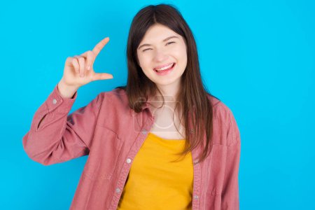 Foto de Joven chica caucásica con camisa rosa aislado sobre fondo azul sonriendo y haciendo un gesto con la mano de tamaño pequeño, símbolo de medida. - Imagen libre de derechos