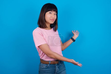 Foto de Joven mujer asiática vistiendo camiseta sobre fondo azul invitando a entrar sonriendo natural con las manos abiertas. Señal de bienvenida. - Imagen libre de derechos