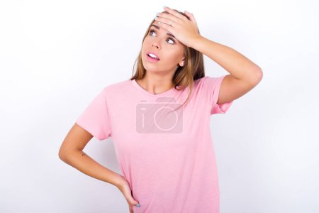 Young Caucasian girl wearing pink T-shirt on white background touching forehead, hears something surprising, glad receive good news, feels relieved. Almost got in trouble.