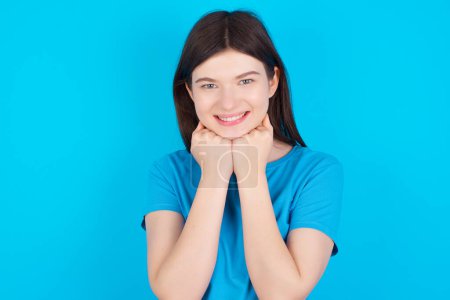 Téléchargez les photos : Satisfait jeune fille caucasienne portant T-shirt bleu isolé sur fond bleu touche menton avec les deux mains, sourit agréablement, se réjouit bonne journée avec amant - en image libre de droit
