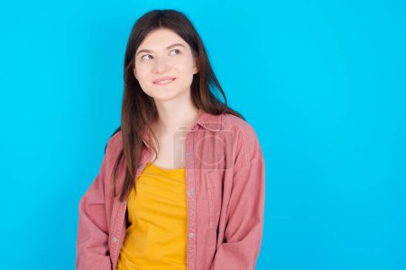 Photo for Young caucasian girl wearing pink shirt isolated over blue background with thoughtful expression, looks away keeps hands down biting his lip thinks about something pleasant. - Royalty Free Image