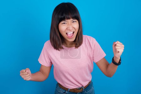 Photo for Portrait of young asian woman wearing t-shirt against blue background looks with excitement at camera, keeps hands raised over head, notices something unexpected reacts on sudden news. - Royalty Free Image