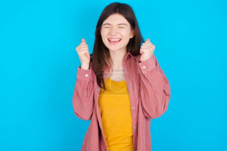 Photo for Young caucasian girl wearing pink shirt isolated over blue background rejoicing his success and victory clenching fists with joy being happy to achieve aim and goals. Positive emotions, feelings. - Royalty Free Image