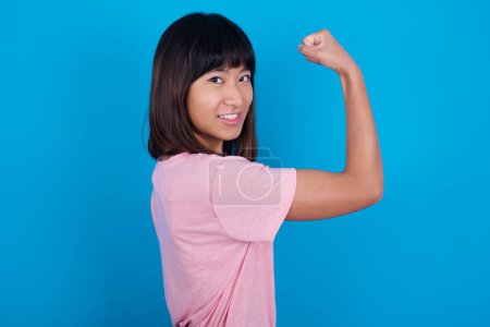 Foto de Joven mujer asiática usando camiseta sobre fondo azul mostrando los músculos después del entrenamiento. Concepto de salud y fuerza. - Imagen libre de derechos