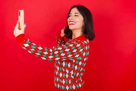 Photo for Brunette caucasian woman wearing christmas sweater over red background smiling and taking a selfie ready to post it on her social media. - Royalty Free Image
