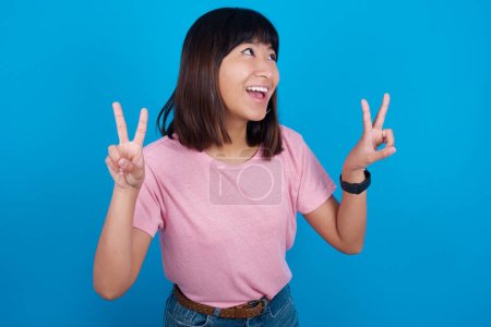 Photo for Isolated shot of cheerful young asian woman wearing t-shirt against blue background makes peace or victory sign with both hands, feels cool. - Royalty Free Image