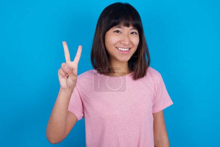 Téléchargez les photos : Jeune femme asiatique portant t-shirt sur fond bleu montrant et pointant vers le haut avec les doigts numéro deux tout en souriant confiant et heureux. - en image libre de droit