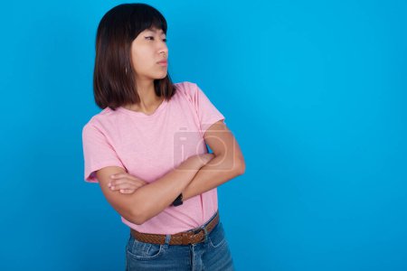 Téléchargez les photos : Image d'une jeune femme asiatique bouleversée portant un t-shirt sur fond bleu avec les bras croisés. Regarder avec expression déçue de côté après avoir écouté les mauvaises nouvelles. - en image libre de droit
