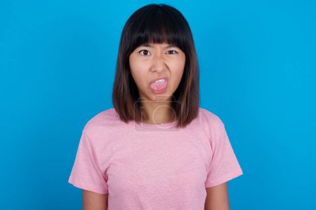 Téléchargez les photos : Langage corporel. Dégoûté stressé jeune femme asiatique portant t-shirt sur fond bleu fronçant les sourcils visage, démontrant une aversion pour quelque chose. - en image libre de droit